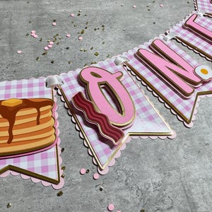 May include: A pink and gold "ONE" banner with a breakfast theme. The banner features pancakes, bacon, and an egg. The letters are pink with gold trim, set against a pink and white gingham pattern.