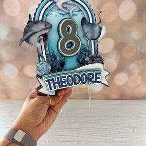 Peut inclure: Un topper de g&acirc;teau bleu et blanc avec un num&eacute;ro 8, un requin, une raie, une tortue et un requin marteau. Le topper indique "Theodore" en lettres bleues.