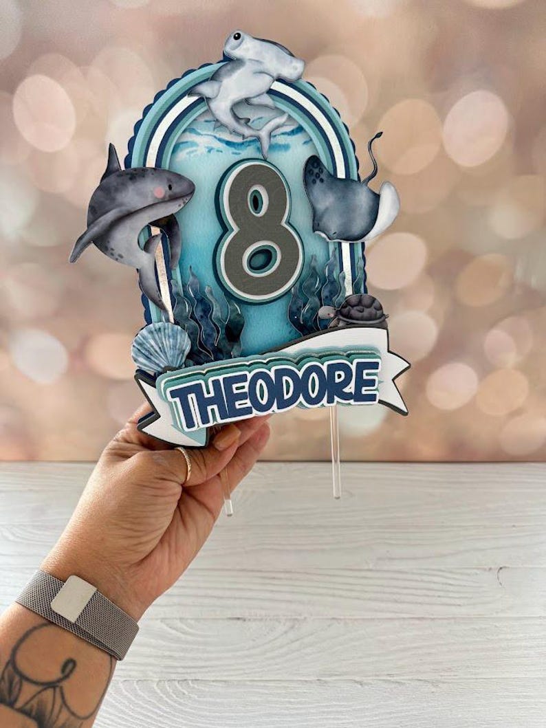 Peut inclure: Un topper de g&acirc;teau bleu et blanc avec un num&eacute;ro 8, un requin, une raie, une tortue et un requin marteau. Le topper indique "Theodore" en lettres bleues.