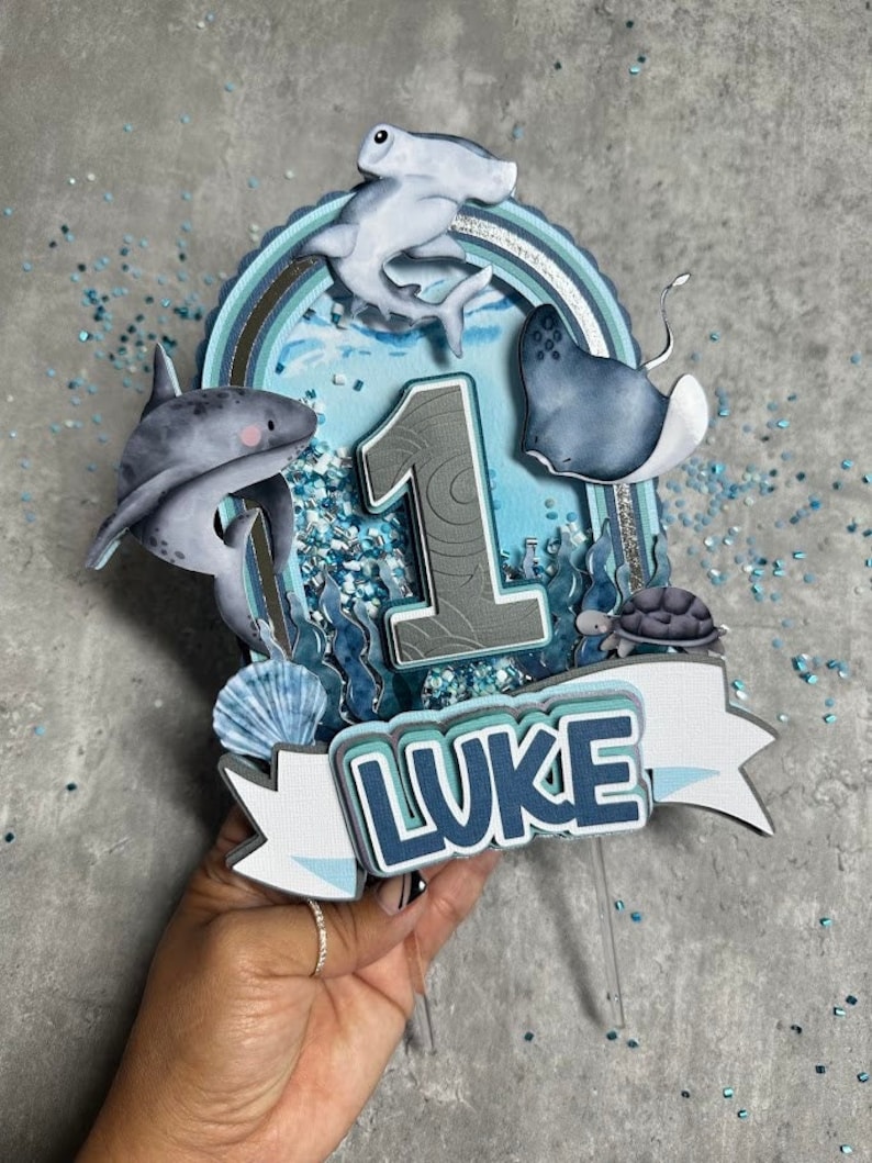 Peut inclure: Un topper de g&acirc;teau bleu et gris avec un num&eacute;ro un, un requin, un requin marteau, une raie, une tortue et le nom "LUKE".