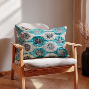 May include: A decorative rectangular pillow with a teal, gray, and cream circular pattern rests on a beige chair. The chair has a light wood frame. The pillow's design features overlapping circles in varying shades of teal, gray, and cream.