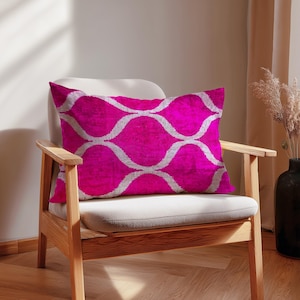 Könnte beinhalten: Ein rechteckiges magenta Kissen mit einem weißen, wellenförmigen Muster liegt auf einem cremefarbenen Stuhl. Das Kissen besteht aus einem weichen Material und setzt einen Farbtupfer in der neutralen Umgebung. Der Stuhl hat einen hellen Holzrahmen.