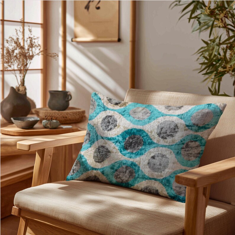May include: A decorative rectangular pillow with a teal, gray, and white circular pattern rests on a light beige chair. The pillow's design features overlapping circles, creating a textured appearance. The chair has a wooden frame.