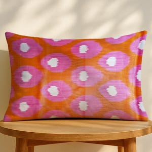 May include: Rectangular decorative pillow with a vibrant orange background and a repeating pattern of pink and white floral-like shapes. The pillow is placed on a wooden table, showcasing its design and color scheme.