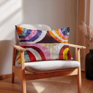 Könnte beinhalten: Ein rechteckiges Zierkissen mit einem lebendigen, abstrakten Design aus konzentrischen Bögen in Rot-, Rosa-, Gelb-, Orange-, Blau- und Lilatönen. Das Kissen liegt auf einem hellen Stuhl mit Holzarmlehnen und -beinen.