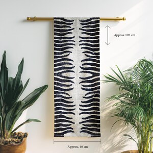 Op de afbeelding: Een rechthoekig, decoratief textiel met een zebraprint, ongeveer 120 cm lang en 40 cm breed, hangt aan een goudkleurige stang. Het zwart-witte ontwerp wordt gepresenteerd tegen een neutrale achtergrond. Potplanten omlijsten het textiel.