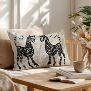 Pode incluir: Almofada decorativa retangular com fundo bege e design de cavalos pretos. Os cavalos estão virados um para o outro e o desenho é feito de pequenos pontos. A almofada está num sofá de cor clara.