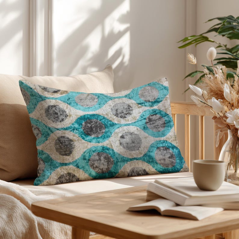 May include: Rectangular decorative pillow with a repeating pattern of teal, gray, and off-white circles. The pillow is placed on a light-colored sofa, with a wooden table and decorative items in the background.