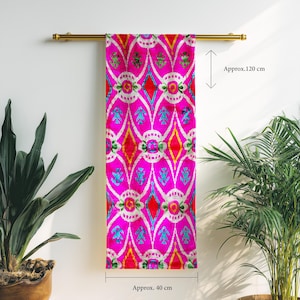 Könnte beinhalten: Ein leuchtend pinkfarbenes Textil mit einem sich wiederholenden Muster aus weißen, roten, gelben und blauen Formen, ca. 40 cm breit und 120 cm lang. Der Stoff hängt an einer goldfarbenen Stange, mit Pflanzen auf beiden Seiten.
