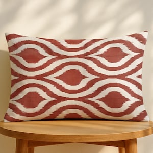 Può includere: Un cuscino rettangolare decorativo con un motivo ripetuto di linee bianche curve su uno sfondo color terracotta. Il cuscino è posto su un tavolo rotondo di legno. Il design ricorda un tessuto ikat.