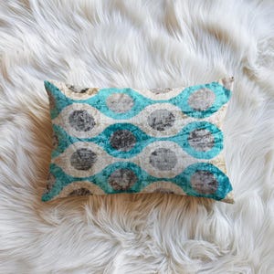 May include: Rectangular decorative pillow with a teal, gray, and cream-colored pattern. The pattern consists of overlapping circles in a wave-like design. The pillow is placed on a white, fluffy surface.