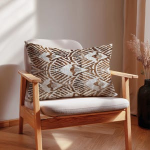 Può includere: Un cuscino rettangolare decorativo con un motivo geometrico marrone e bianco poggia su una sedia beige. Il design del cuscino presenta forme curve e lineari ripetute in varie tonalità di marrone su uno sfondo bianco. La sedia ha una struttura in legno.