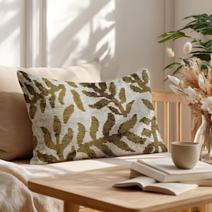 May include: Rectangular decorative pillow with a botanical pattern in olive green and brown on a cream background. The pillow is placed on a light-colored sofa. The scene includes a wooden table, books, and a vase with dried flowers.