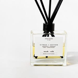 May include: A clear square glass reed diffuser bottle with black reeds. The white label reads: HAND POURED N° 001 CITRUS + COTTON GRAPEFRUIT • MANDARIN LINEN • SANDALWOOD wash + wik REED DIFFUSER. The bottle contains a pale yellow liquid fragrance oil.