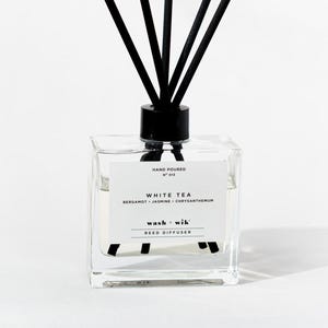 May include: A clear square glass reed diffuser bottle with a black cap and five black reeds. The white label reads: HAND POURED, NO 012, WHITE TEA, BERGAMOT + JASMINE + CHRYSANTHEMUM, wash + wik, REED DIFFUSER. The bottle contains a clear liquid.