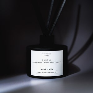 May include: A black diffuser bottle with a white label that reads "HAND POURED N° 043 SANTAL SANDALWOOD MUSK AMBER CEDAR wash + wik SMALL BATCH // ORLANDO, FL".