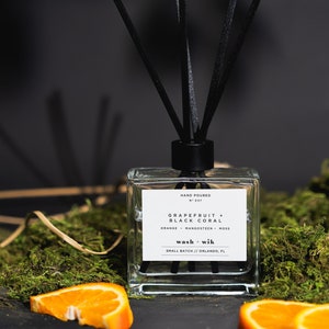 May include: A clear glass diffuser bottle with black reeds, labeled "Grapefruit + Black Coral." The label lists orange, mangosteen, and moss. The bottle sits on a bed of green moss with orange slices. The text "wash + wik" and "Small Batch // Orlando, FL" are also visible.