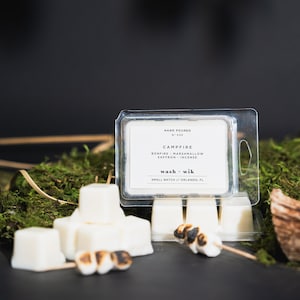 May include: A white wax melt in a clear plastic container with the text "HAND POURED M030 CAMPFIRE BONFIRE MARSHMALLOW SAFFRON INCENSE wash wik SMALL BATCH//ORLANDO, FL". The wax melt is surrounded by white wax melts and a few toasted marshmallows on a wooden stick.
