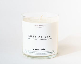 May include: A clear glass jar candle with a white label featuring the text "HAND POURED N° 010", "LOST AT SEA", "OZONE • SEA SALT • OAKMOSS • CITRUS", "wash + wik", and "SMALL BATCH // 100% SOY WAX CANDLE // ORLANDO, FL". The candle has a single cotton wick.