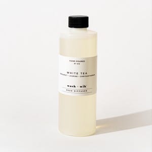 May include: A clear plastic bottle filled with light yellow liquid, topped with a black screw cap. The white label reads: HAND POURED NO 013, WHITE TEA, BERGAMOT JASMINE CHRYSANTHEMUM, wash + wik, REED DIFFUSER. The bottle has a cylindrical shape.