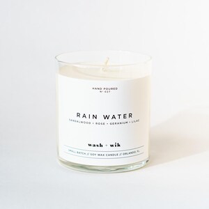 Op de afbeelding: Een kaars in helder glas met een wit etiket met de tekst RAIN WATER, SANDALWOOD, ROSE, GERANIUM, LILAC en wash + wik. Het etiket bevat ook HAND POURED N 027 en SMALL BATCH // SOY WAX CANDLE // ORLANDO, FL. De kaars heeft een enkele lont.