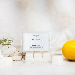 May include: A clear plastic container holding white wax melts. The label reads "NO SQUITO" with ingredients listed. A whole yellow lemon is to the right. The product is from "wash + wik" in Orlando, FL.