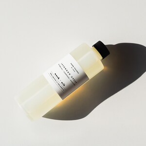 May include: A clear rectangular bottle with a black cap, filled with a light yellow liquid. The bottle has a white label with black text that reads "Lavender Dream" and "wash + wik". The bottle is angled on a white surface.