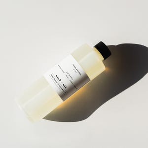 May include: A rectangular, clear bottle with a black cap, containing a light yellow liquid. The bottle has a white label with black text that reads "SANTAL" and "wash + wik". The product is hand-poured and made in Orlando.