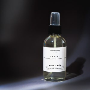May include: A clear glass bottle with a black spray top and a white label. The label reads "HAND POURED N° 043 SANTAL SANDALWOOD MUSK AMBER CEDAR wash + wik SMALL BATCH // ORLANDO, FL"
