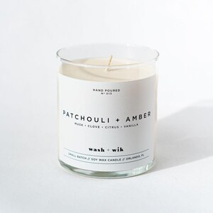 Puede incluir: Una vela de cera de soja color crema en un frasco de vidrio transparente con una mecha central. La etiqueta blanca presenta el texto negro: Hand Poured No 015, Patchouli + Amber, Musk, Clove, Citrus, Vanilla, wash + wik, Small Batch // Soy Wax Candle // Orlando, FL.
