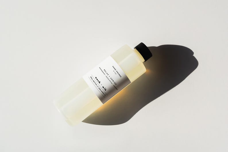May include: A rectangular, clear bottle with a black cap, filled with a light yellow liquid. A white label with black text reads "Relax + Unwind" and "wash + wik". The bottle casts a long shadow on a white surface.