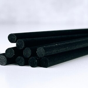 May include: A close-up shot of several black diffuser sticks. The sticks are cylindrical, with a slightly textured surface. They are arranged in a loose bundle, with some sticks overlapping. The background is a plain white.