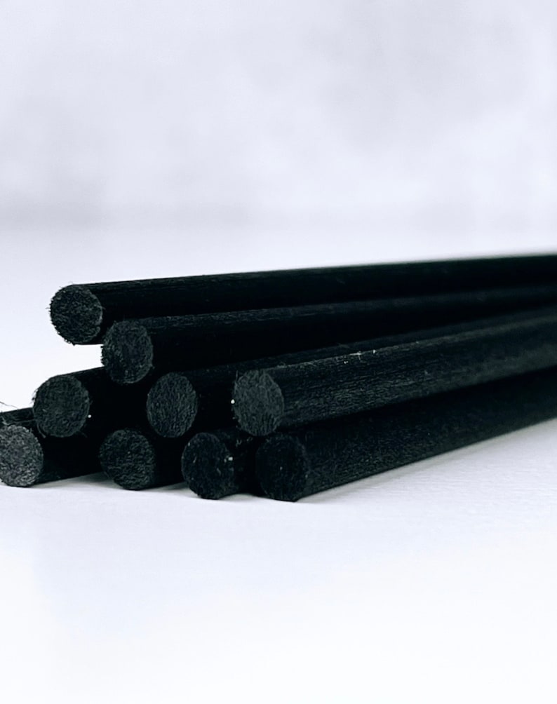 May include: A close-up shot of several black diffuser sticks. The sticks are cylindrical, with a slightly textured surface. They are arranged in a loose bundle, with some sticks overlapping. The background is a plain white.