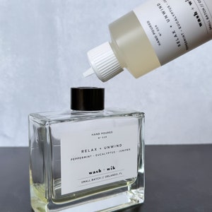 May include: A clear, square glass bottle with a black cap and a white label that reads "RELAX + UNWIND" and "wash + wik." A second bottle is pouring liquid into the first. The label also says "Peppermint, Eucalyptus, Juniper."