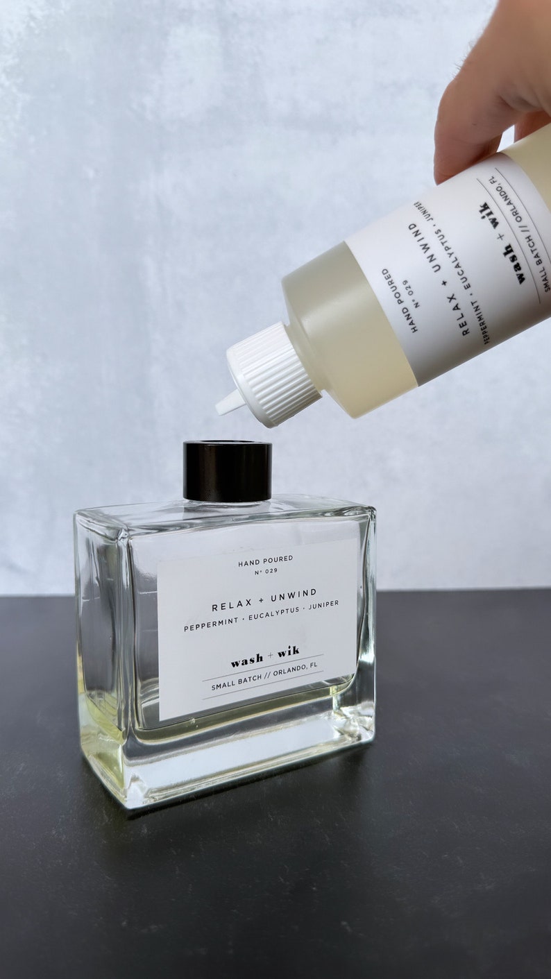 May include: A clear, square glass bottle with a black cap and a white label that reads "RELAX + UNWIND" and "wash + wik." A second bottle is pouring liquid into the first. The label also says "Peppermint, Eucalyptus, Juniper."