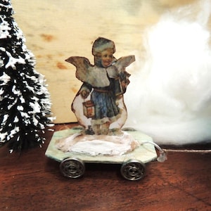 Könnte beinhalten: Eine Miniatur-Weihnachtsengel-Figur auf einer Plattform mit Rädern. Der Engel trägt blaue und weiße Kleidung und hält eine Laterne. Ein kleiner, schneebedeckter Weihnachtsbaum befindet sich links vom Engel. Der Hintergrund umfasst weißes, flauschiges Material.