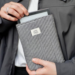 May include: A black and white gingham e-reader case with the text "aeny bags" on a white label. The case is being held by a person wearing a gray jacket.