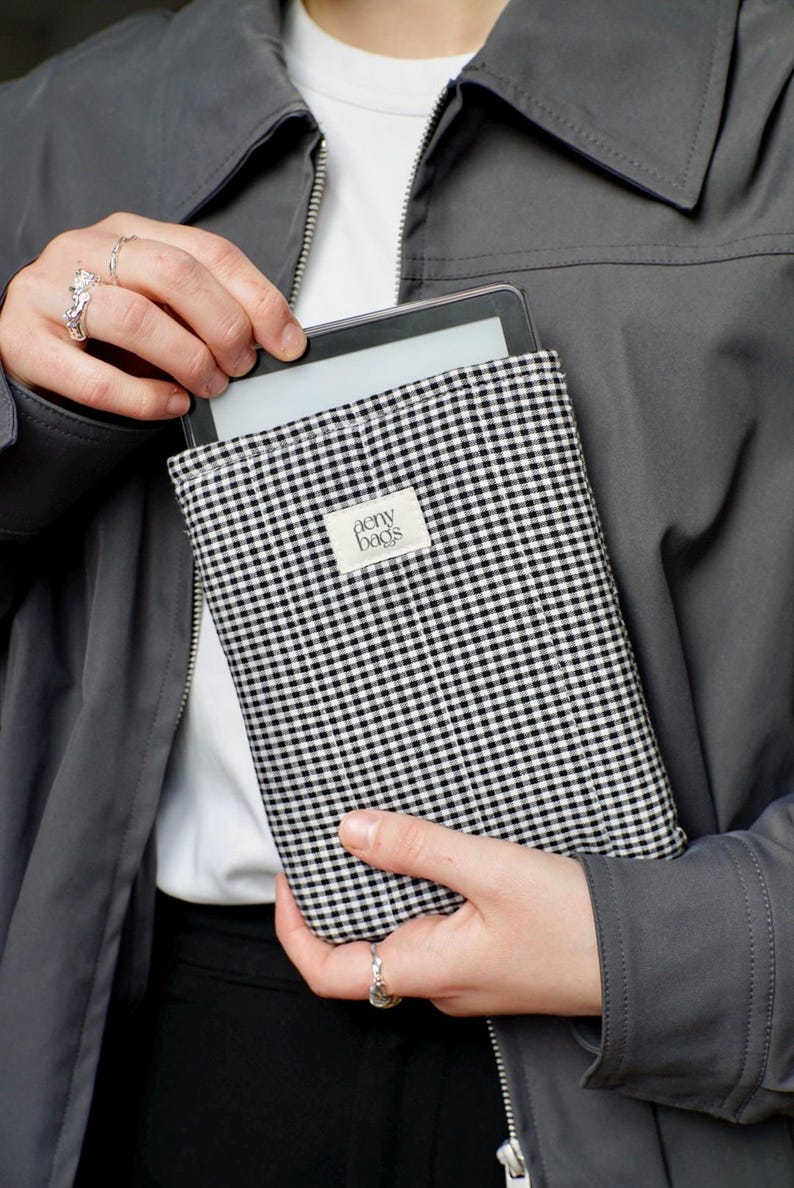 May include: A black and white gingham e-reader case with the text "aeny bags" on a white label. The case is being held by a person wearing a gray jacket.
