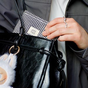 May include: A black leather tote bag with a white and black checkered lining. The bag has a gold ring and a white fluffy keychain. The lining has a white label that says "penny bags".