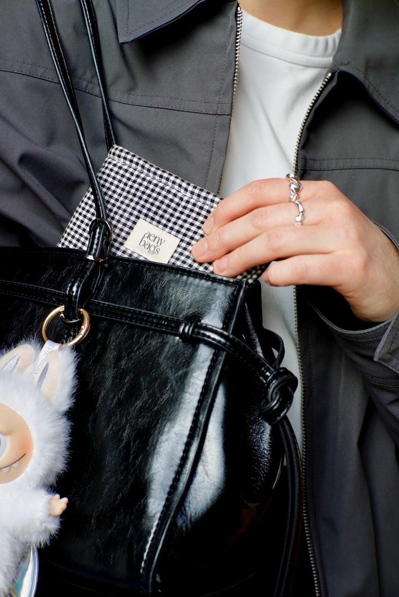 May include: A black leather tote bag with a white and black checkered lining. The bag has a gold ring and a white fluffy keychain. The lining has a white label that says "penny bags".