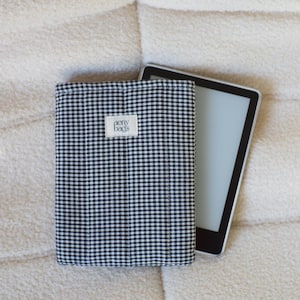 May include: A black and white gingham e-reader case with the text "aeny bags" on the front. The case is lying on a white fluffy surface.