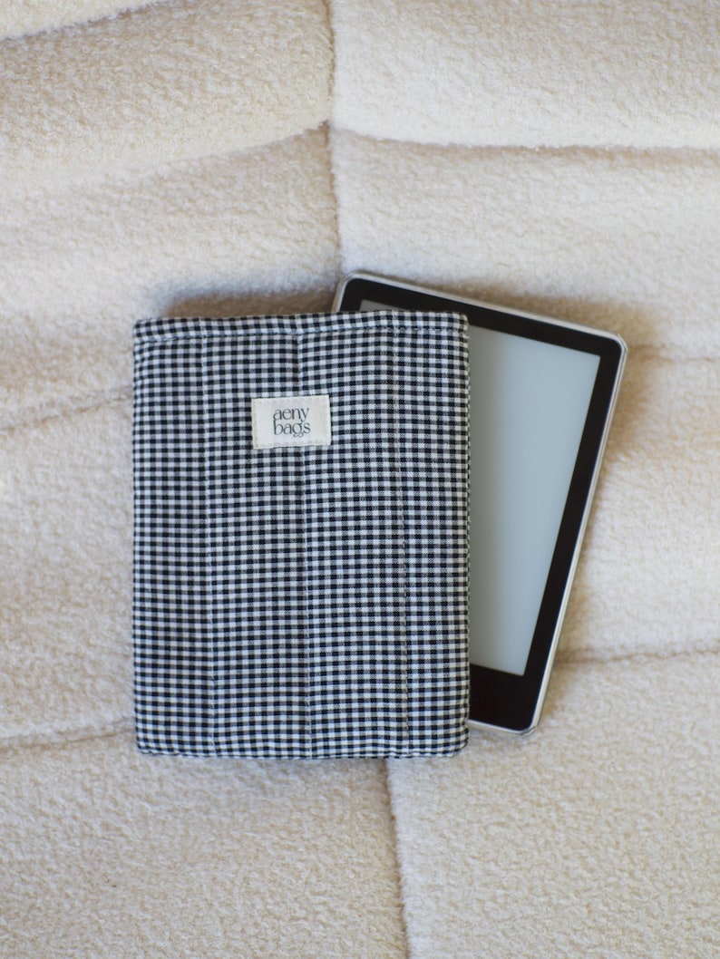 May include: A black and white gingham e-reader case with the text "aeny bags" on the front. The case is lying on a white fluffy surface.