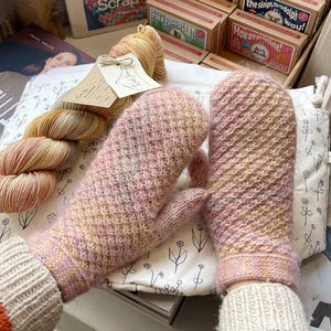 May include: A pair of pink and yellow knitted mittens. The mittens are made of a soft, textured yarn and have a ribbed cuff. A skein of yarn and a book are visible in the background. The mittens are a cozy accessory.