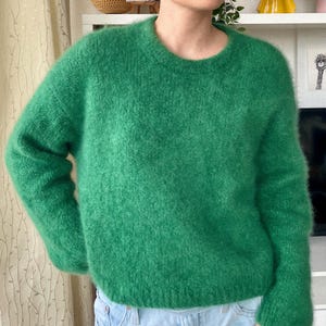 Puede incluir: Un suéter verde brillante con cuello redondo. El suéter tiene una textura suave y difusa y mangas largas. El modelo lleva vaqueros azul claro. El suéter es de color liso y parece estar hecho de un material cálido.