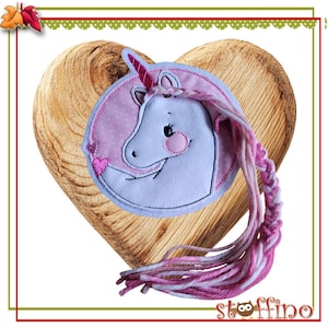 Könnte beinhalten: Ein rosa-weißes Einhorn-Applikationen mit einem rosa Herzen und einem langen rosa-weißen geflochtenen Schwanz. Die Applikation befindet sich auf einem rosa Hintergrund mit weißen Punkten.