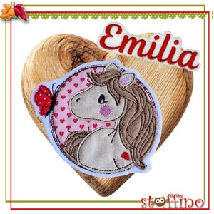 Könnte beinhalten: Eine herzförmige Holzplakette mit einem rosa-weißen gestickten Aufnäher, der ein braunes Pferd mit einem Schmetterling zeigt. Der Name "Emilia" ist auf einem weißen Aufnäher über dem Pferd gestickt.
