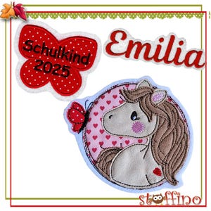 Könnte beinhalten: Drei Bügelbilder für Kleidung. Ein roter Schmetterling mit weißen Punkten und dem Text "Schulkind 2025". Ein weißes Bügelbild mit dem Namen "Emilia" in roten Buchstaben. Ein rundes Bügelbild mit rosa Hintergrund und weißen Herzen, das ein braunes und graues Pony mit rosa Nase zeigt.