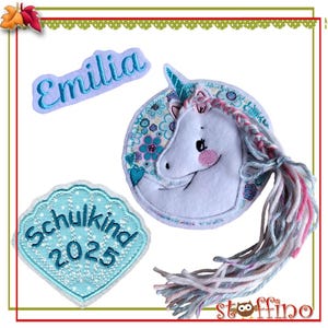 Könnte beinhalten: Drei gestickte Patches für einen Kinderrucksack oder Kleidung. Die Patches sind blau, weiß und rosa. Ein Patch sagt "Emilia" in blauer Schrift. Ein anderer Patch sagt "Schulkind 2025" in blauer Schrift. Der dritte Patch ist ein weißes Einhorn mit einer rosa und blauen Mähne und einem Schwanz.