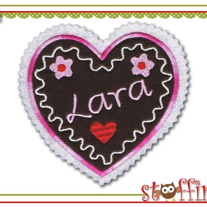 Könnte beinhalten: Ein herzförmiger Aufnäher mit dem Namen "Lara" in Rosa gestickt. Der Aufnäher hat eine dunkelbraune Mitte, einen rosa Rand und einen weißen, gewellten Rand. Gestickte Blumen und ein rotes Herz sind ebenfalls vorhanden.