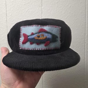 Peut inclure: Casquette de baseball en velours côtelé noir avec un écusson rectangulaire représentant un poisson coloré. Le poisson a des nageoires rouges, un corps bleu et se détache sur un fond bleu clair. L'écusson est cousu sur la casquette.